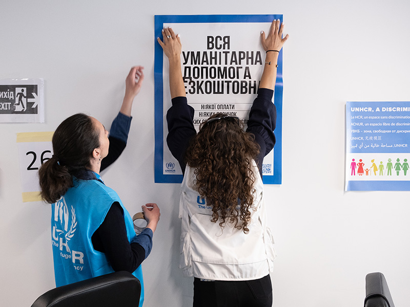 wo UNHCR staff are hanging a poster on a wall of the cash registration center