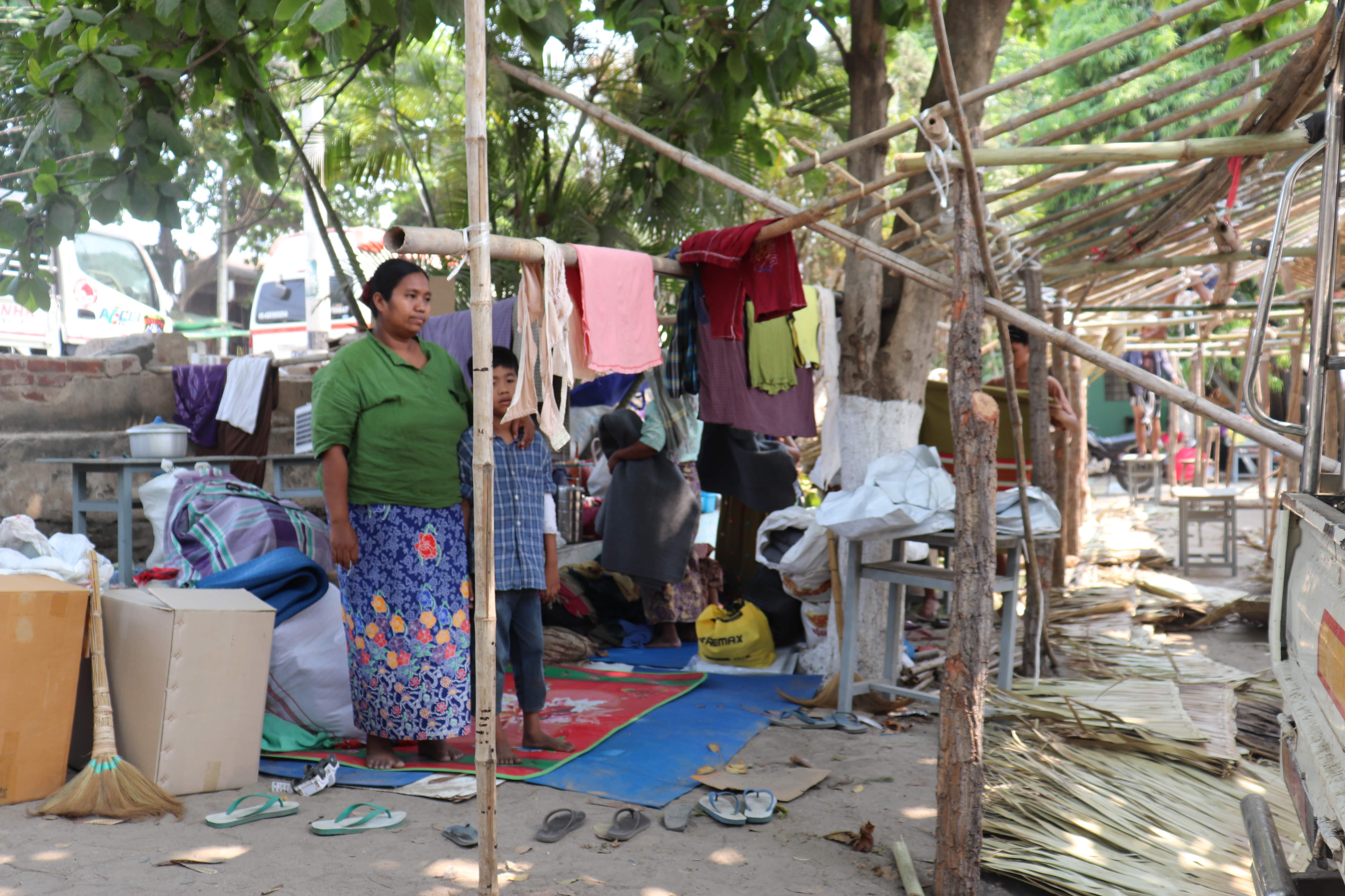 A woman stands under a tree with her arm around a young boy next to an unfinished bamboo structure and surrounded by belongings