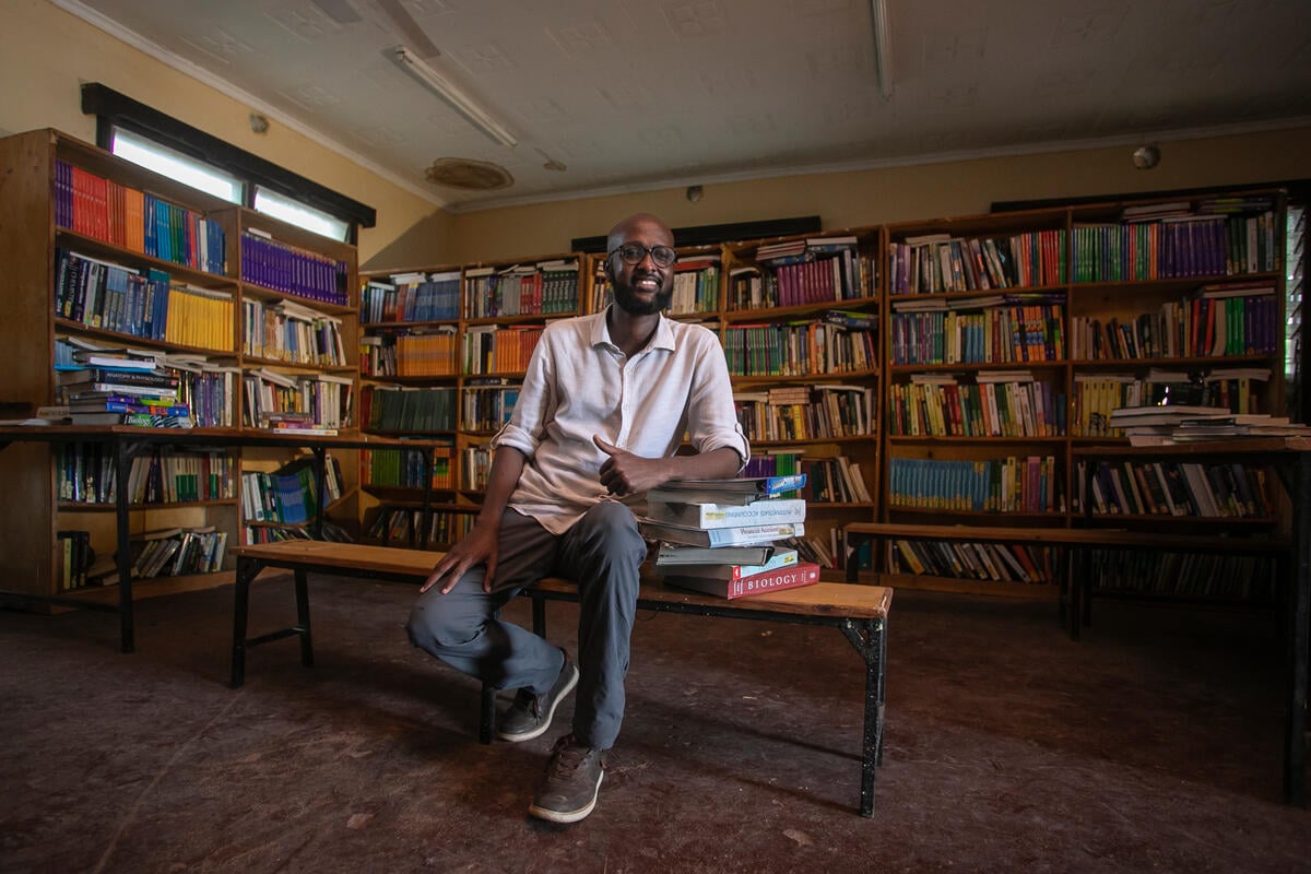 A man sits in a library, surrounded by books and leaning on a pile of books