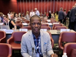 A man sits in a conference room with other delegates in the background.