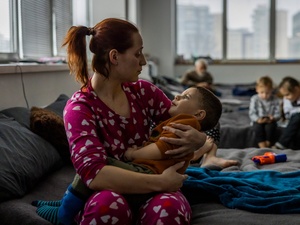 A woman sits on a camp bed holding her young son in a collective shelter for refugees.