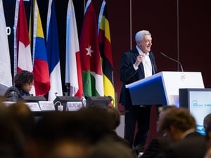 A man in a suit gestures while speaking at a lectern in front of a crowd
