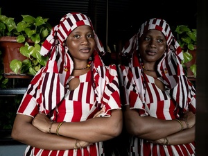 A woman in a red and white striped dress and headscarf leans against a window in which she is reflected