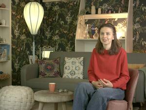 A woman with long brown hair and a red shirt sits in a living room with floral wallpaper.