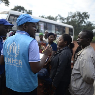 A UNHCR staff member writes on a clipboard while men and women queue for a bus.