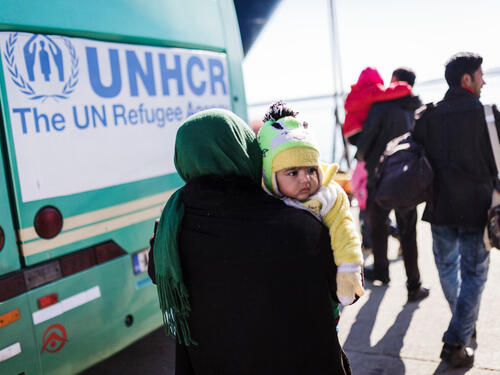 A woman carrying her young child walks towards a UNHCR bus.