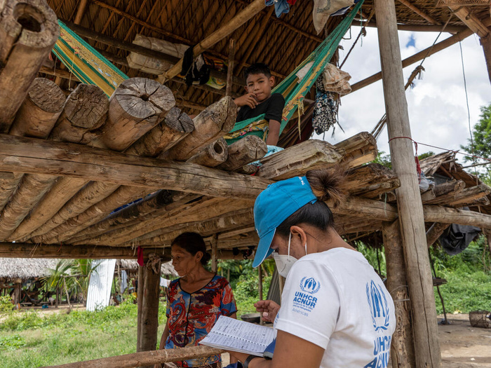 A UNHCR staff member writes on a notebook in front of a woman, and a young boy sitting on a hammock.