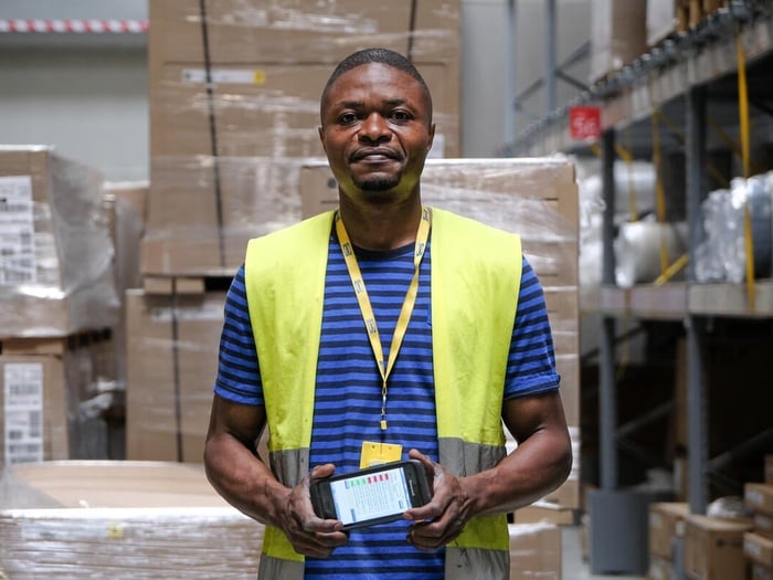 A man wearing an IKEA lanyard stands in a warehouse, holding a mobile device and smiling at the camera.