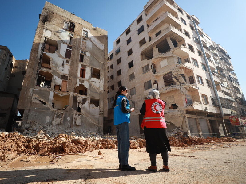 A UNHCR staff member and Red Cross staff member stand in front of a destroyed building.