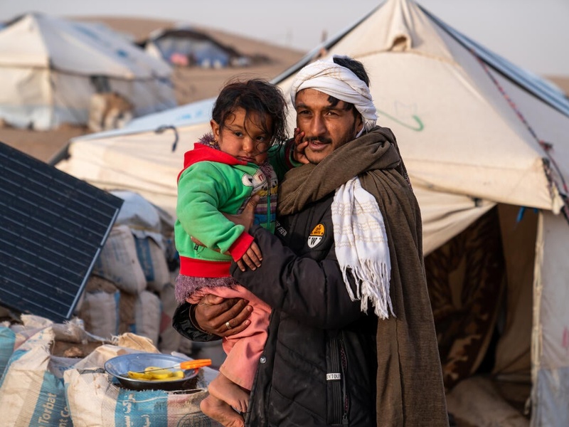 An internally displaced man in Afghanistan, wrapped in blankets, holds a small child. Behind him is a refugee tent settlement.