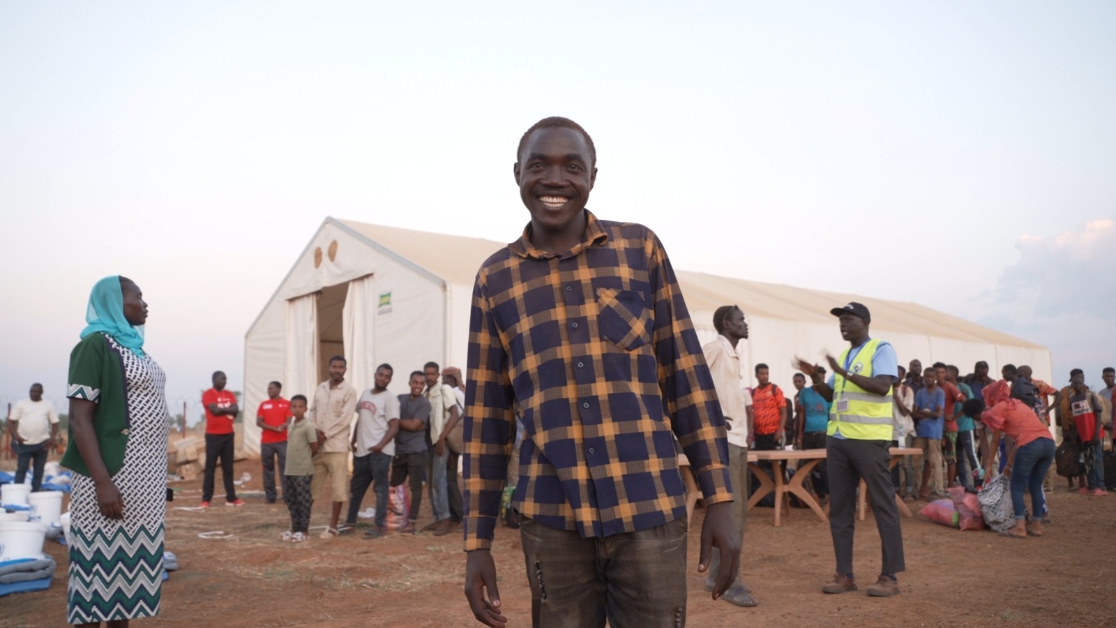 A smiling man stands in front of a large tent where people are standing in line.