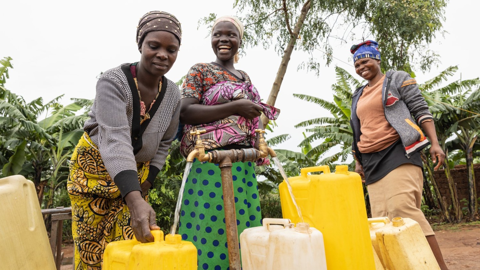 A woman fills up a container at a tap while two other women watch on.