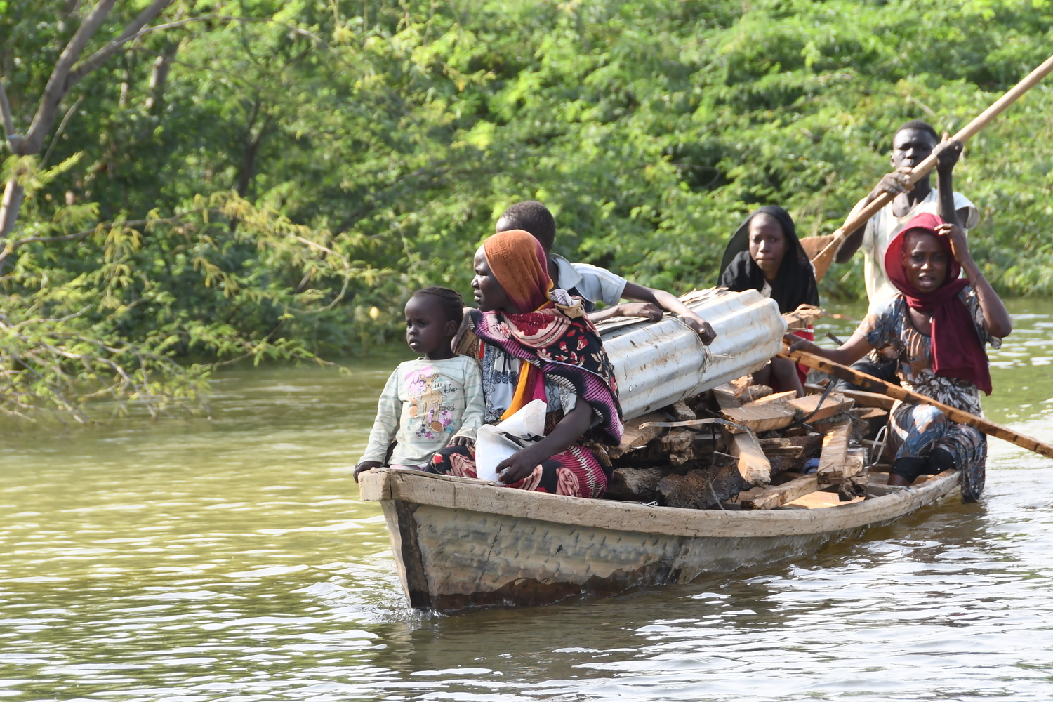 Several adults and children travel in a dugout canoe piled with timber and corrugated iron.