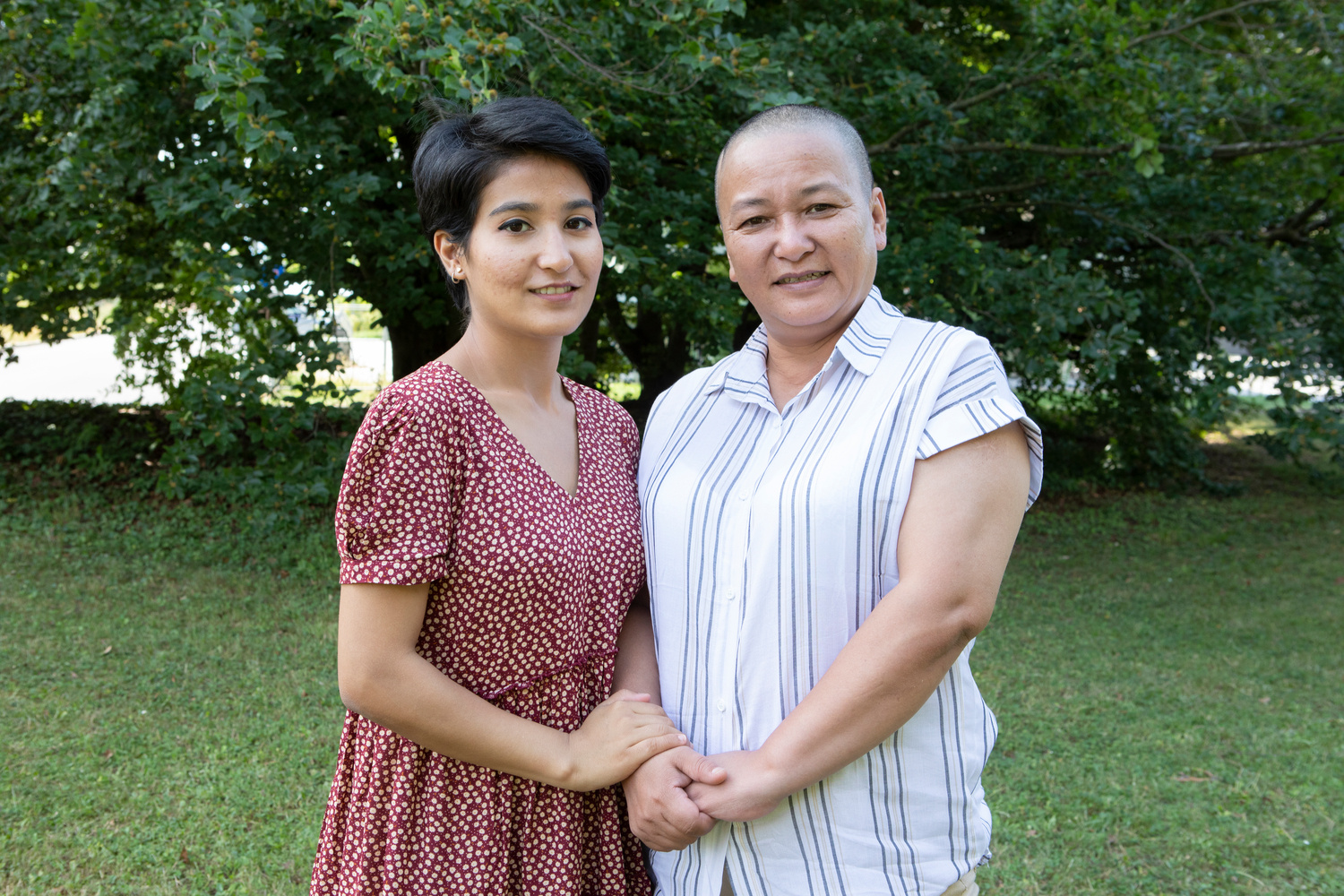A mother and daughter stand together on a law backed by trees.