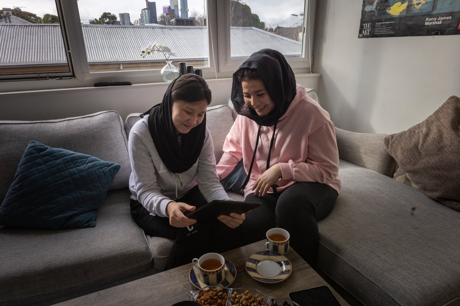 Two young women sit on a couch together looking at a tablet and drinking tea.