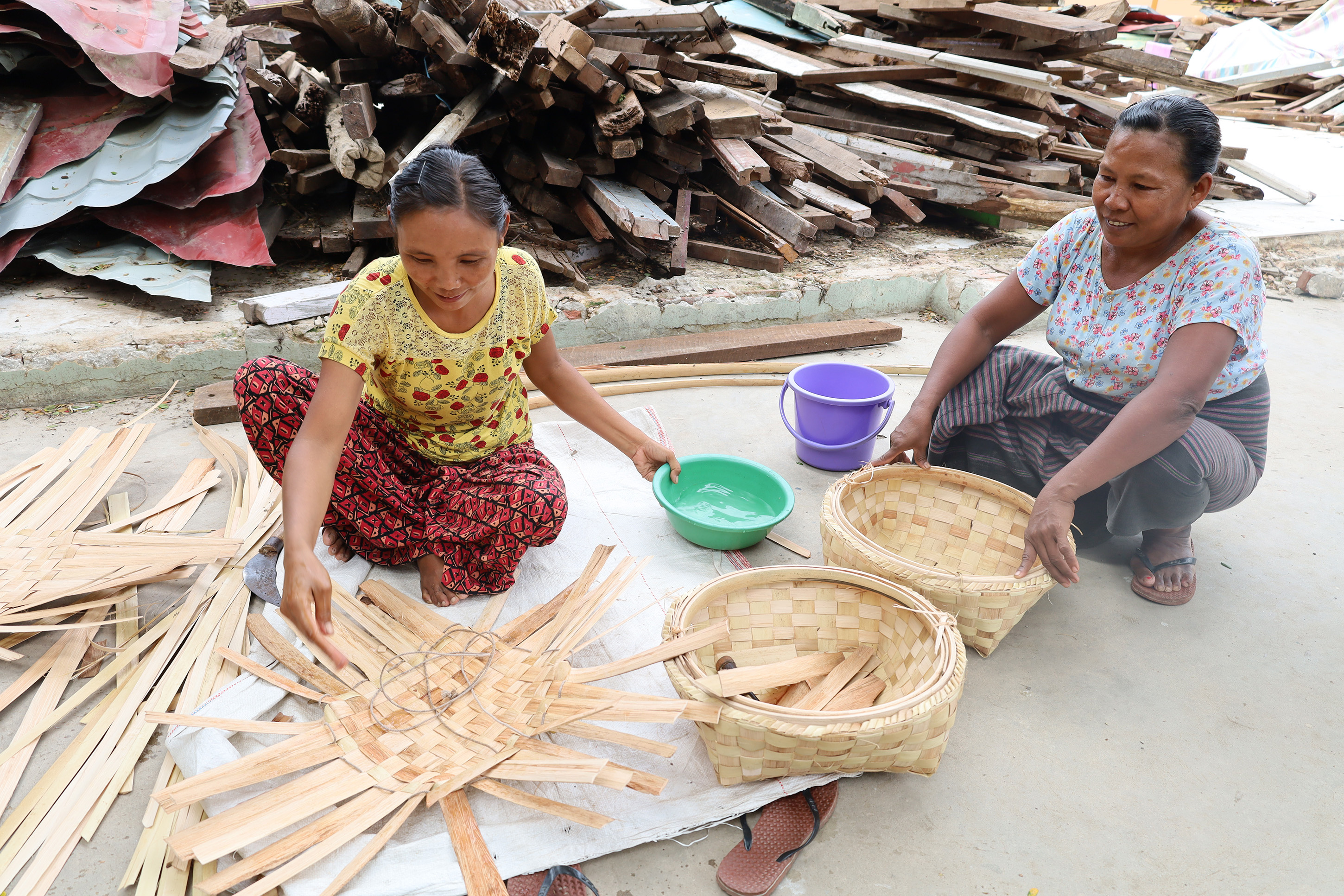 Daw Swe Win weaves baskets in front of a pile of wood – the remains from her shelter destroyed by the earthquake