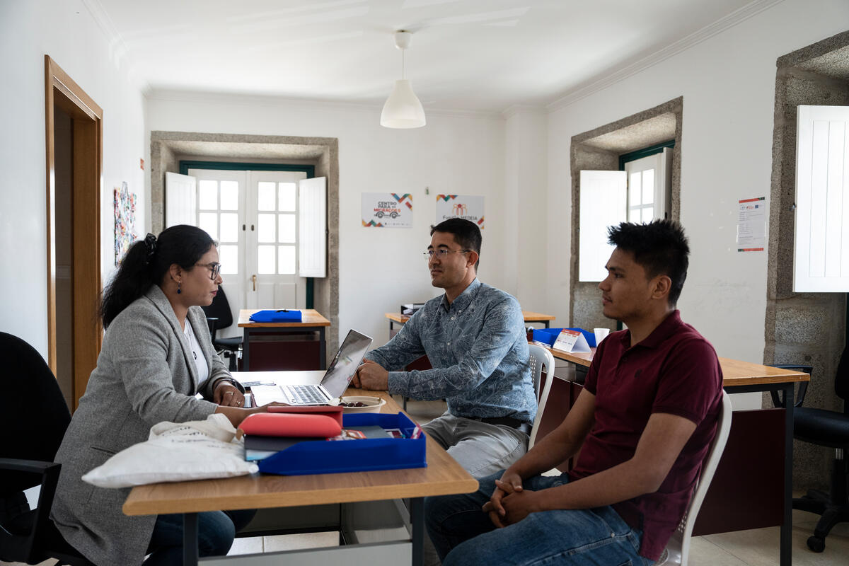 A woman seated behind a desk speaks to two men in her office.