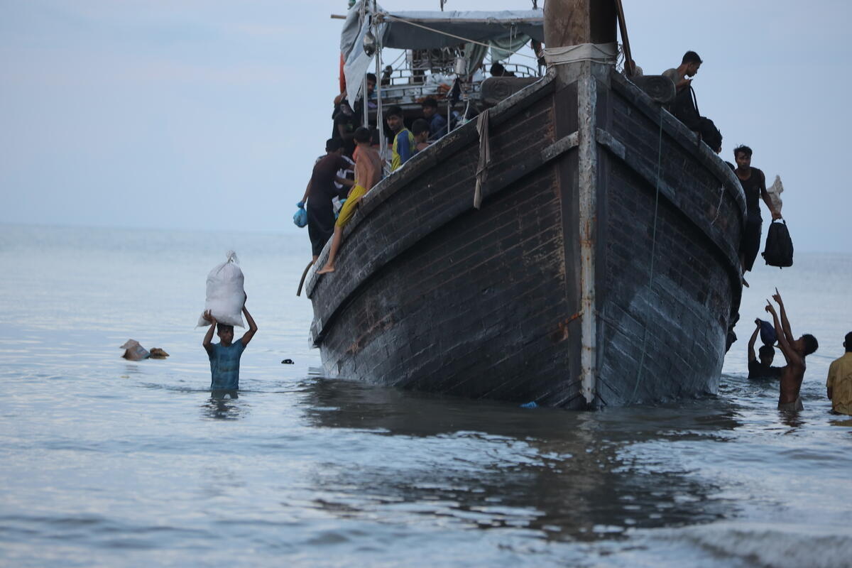 Passengers unload belongings from a wooden boat to people standing in the shallows.