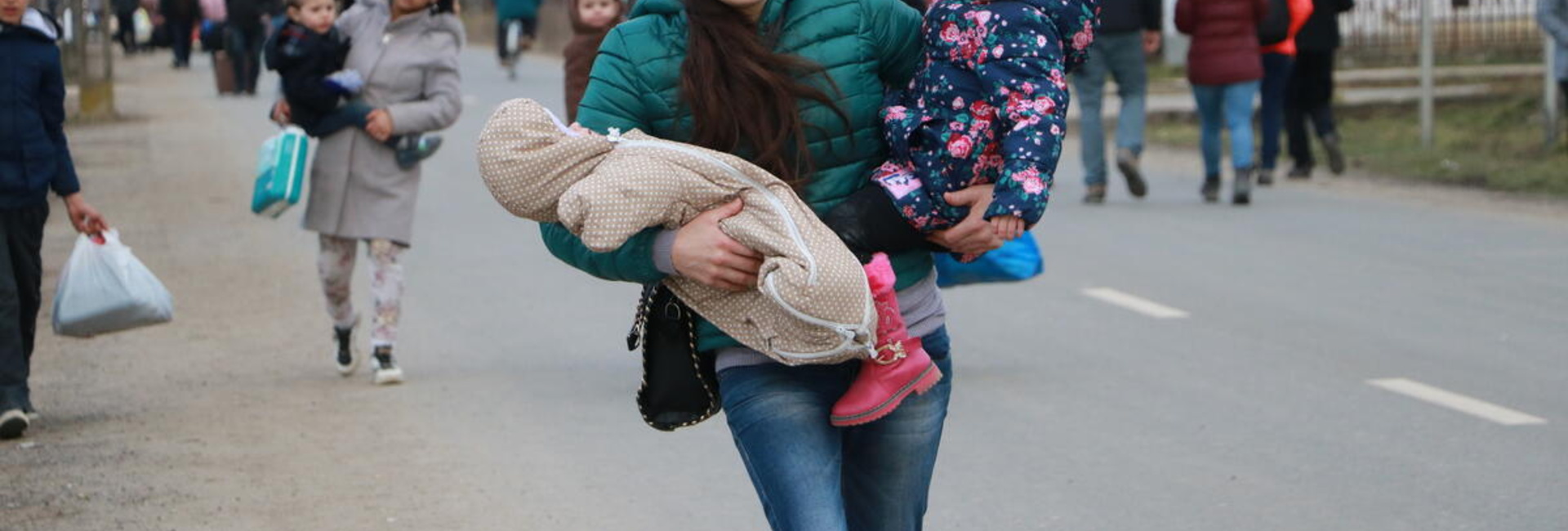 A young woman walks along the side of a road carrying a toddler and a baby in her arms 