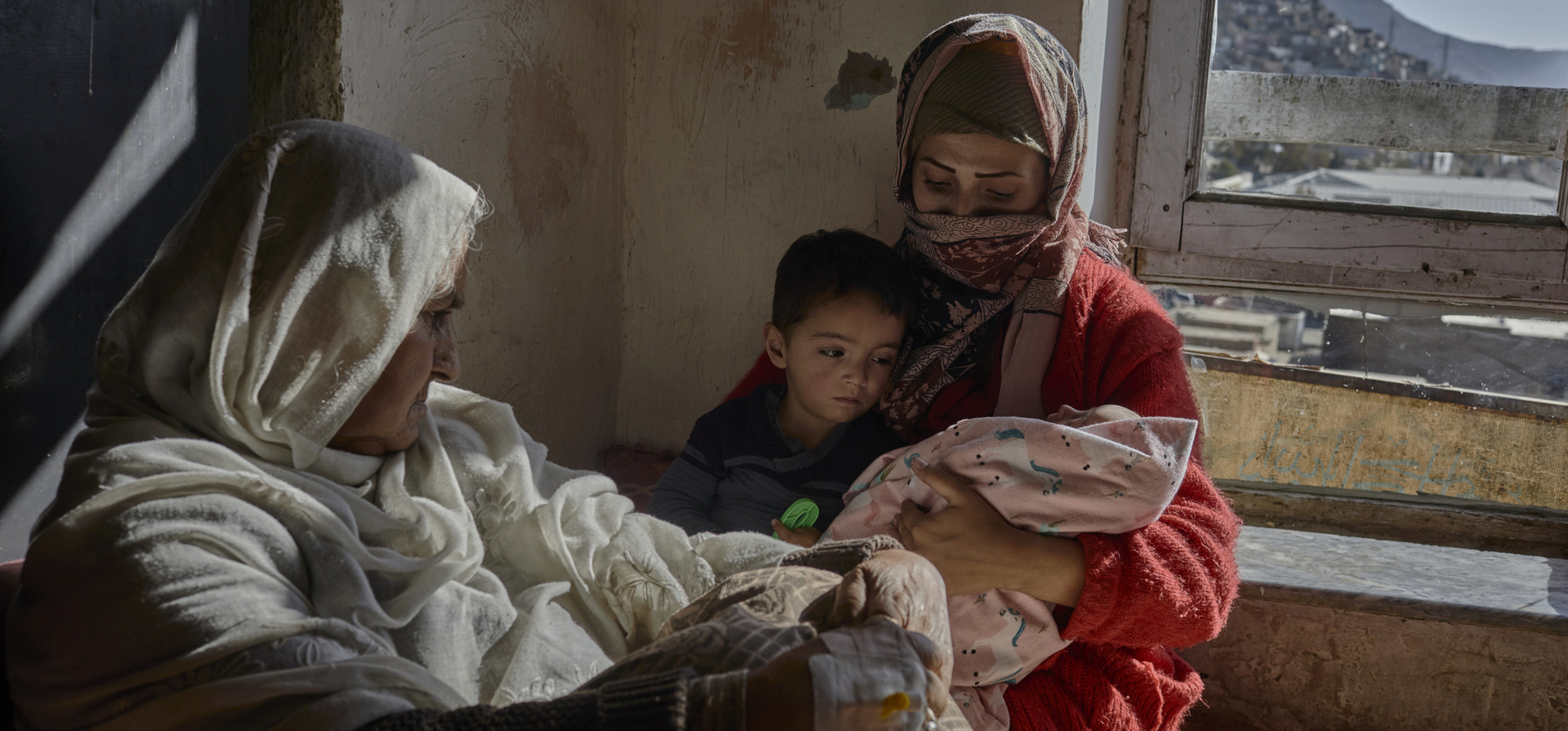 A mother cradles her baby in a bare room with her son and mother looking on