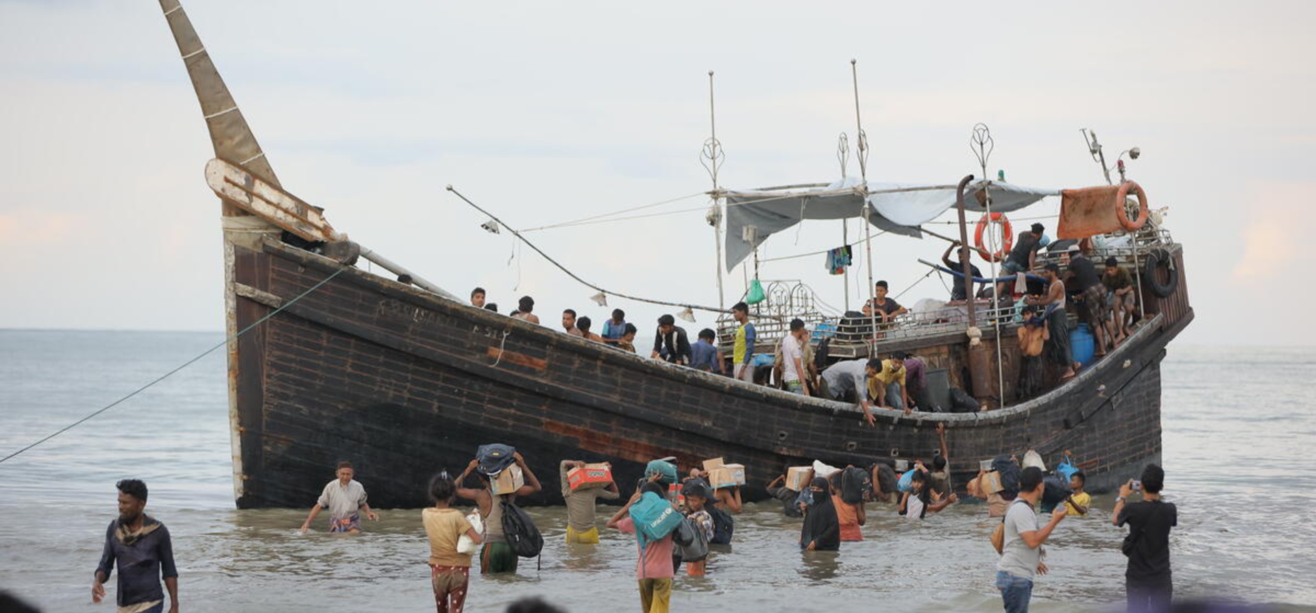 A group of people carrying belongings stand in the shallows alongside a wooden boat carrying refugees.