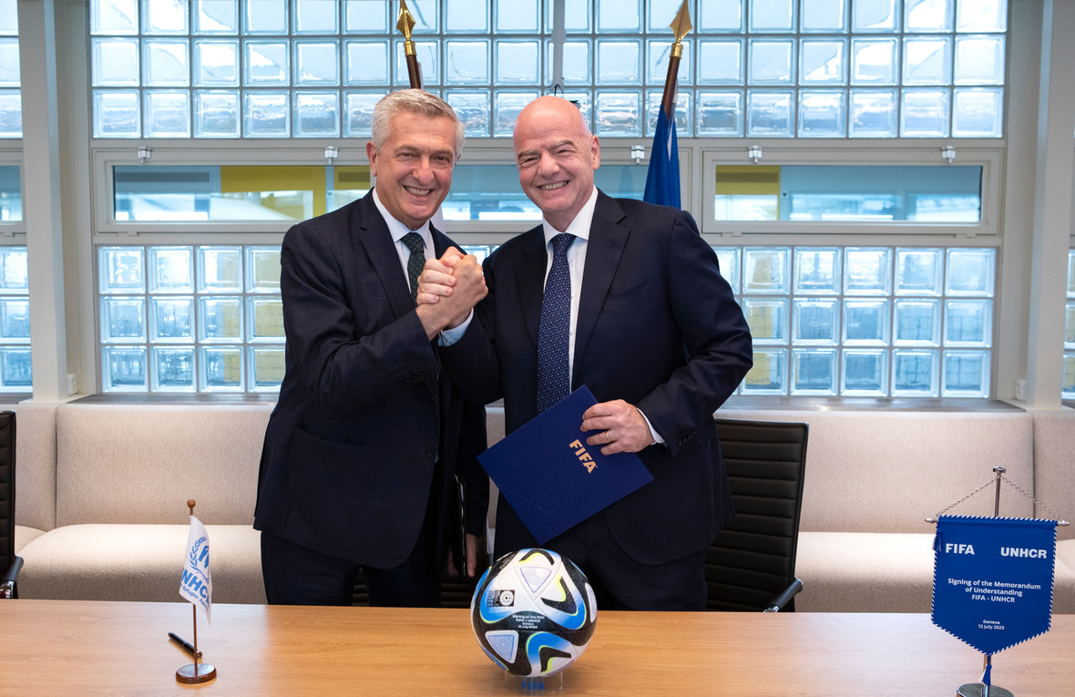Two men shake hands behind a desk with a football on it.