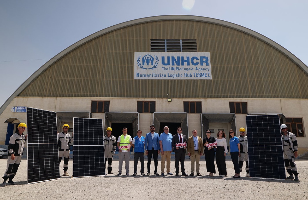 Fourteen staff from UNHCR and LONGi stand outside a large logistics hub, holding up several solar panels