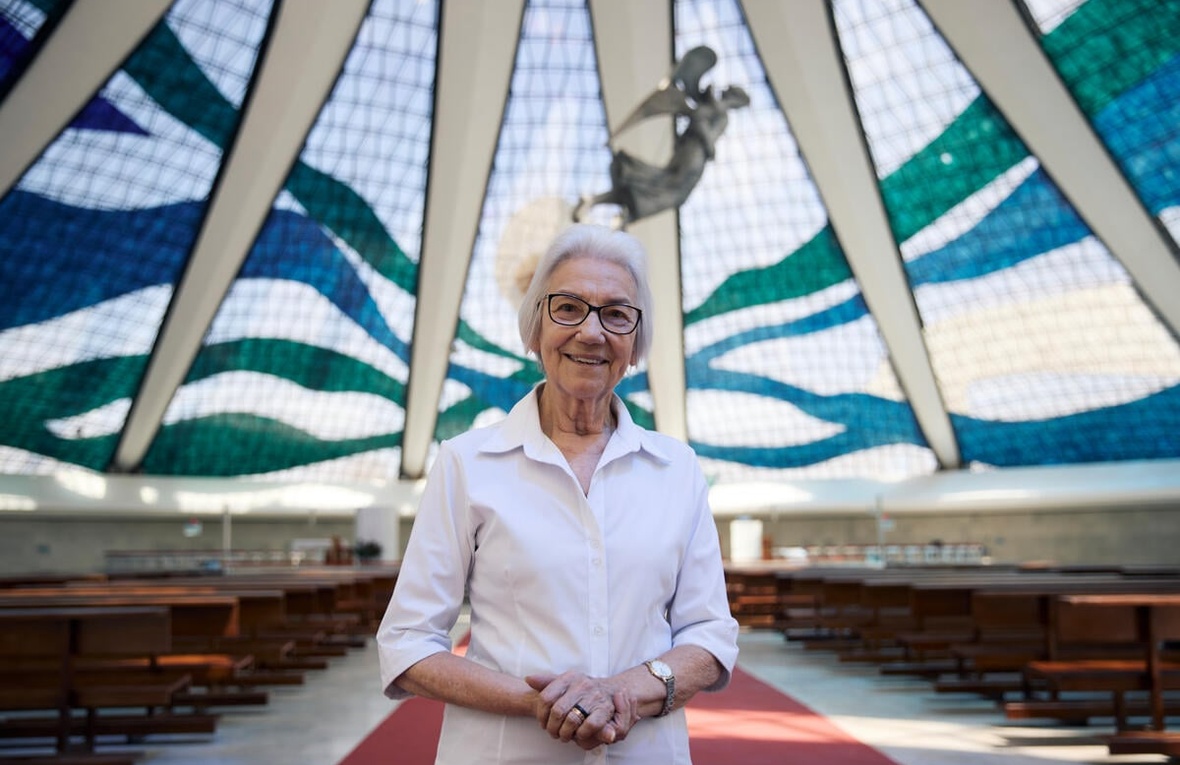 A woman with white hair weaing glasses and a white shirt stands inside a large glass-domed church.