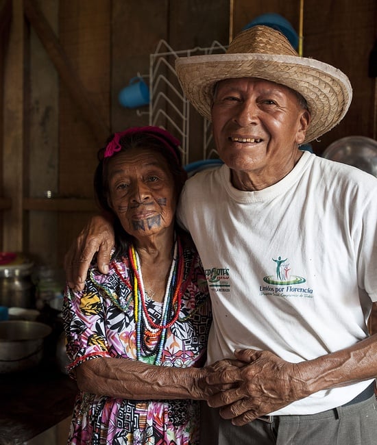 An old couple hold hands and smile at the camera.