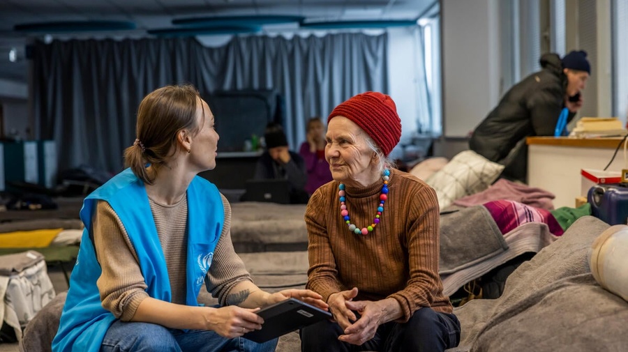 A UNHCR staff member speaks with a Ukrainian refugee inside a temporary shelter, a large hall filled with rows of beds.