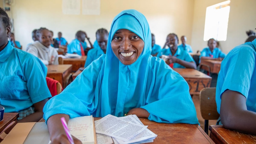 A young woman wearing a headscarf sits at a desk in a classroon.