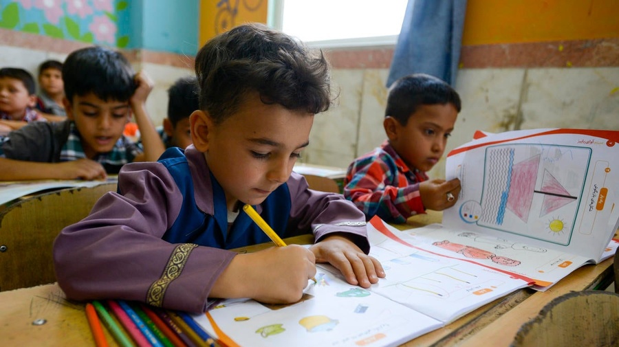 Un niño escribiendo en un cuaderno; a su alrededor, otros niños en un aula