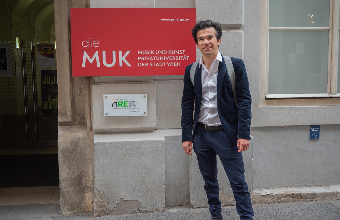 Junger Mann mit dunklen Haaren steht vor Geb&auml;ude mit Schild