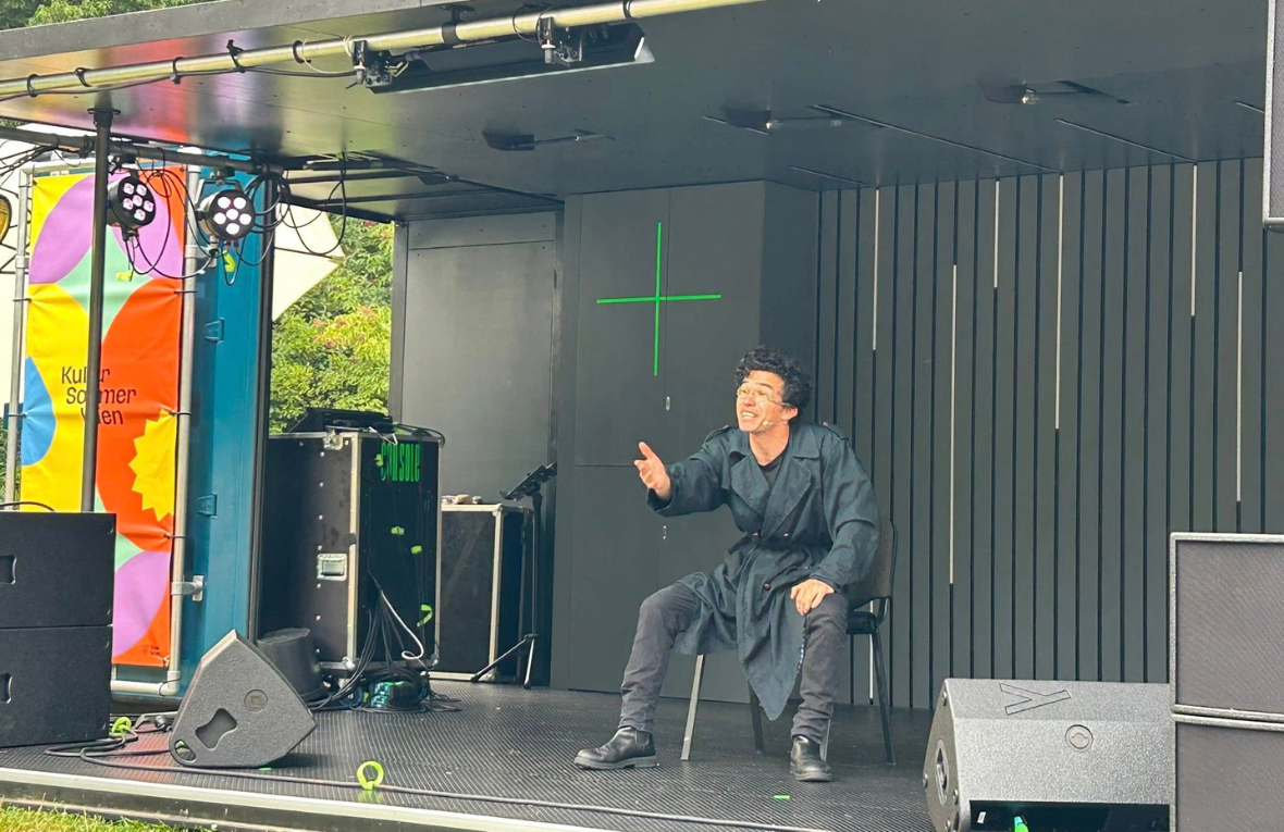 Mann mit Brille und dunkler Kleidung auf einer Theaterb&uuml;hne im Freien.