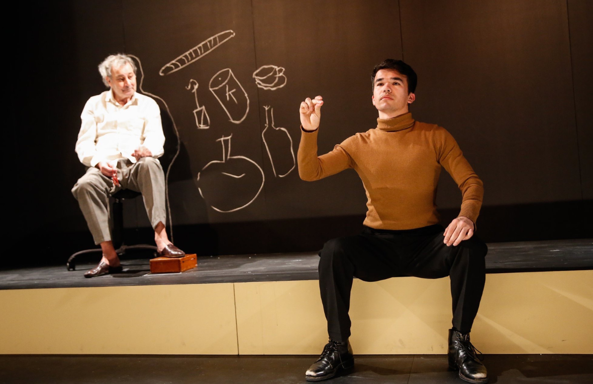 Zwei M&auml;nner sitzen auf einer Theaterb&uuml;hne mit schwarzer Tafelwand und Kreidezeichnungen im Hintergrund