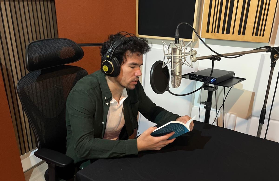 Mann in Tonstudio mit Kopfh&ouml;rern sitzt vor einem Mikrofon und liest aus einem Buch