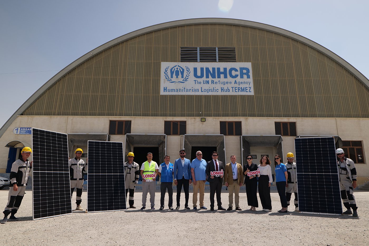 Fourteen staff from UNHCR and LONGi stand outside a large logistics hub, holding up several solar panels