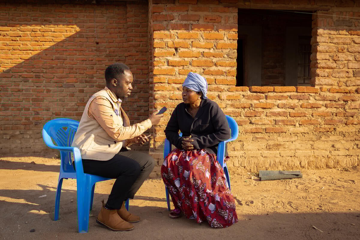 A man interviews a woman outside on two plastic chairs, in front of a brick building