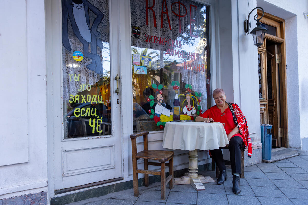A smiling woman seated at a table in front of a café with colorful paintings on the windows and doors.