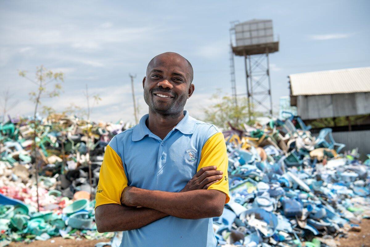 A smiling man stands in front of a huge pile of plastic waste.
