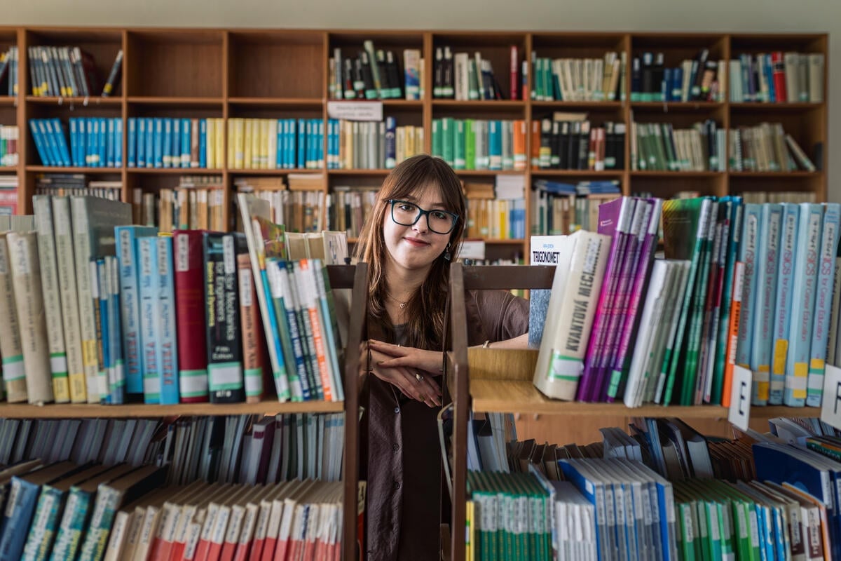 A woman in glasses smiles from between bookshelves in a library.