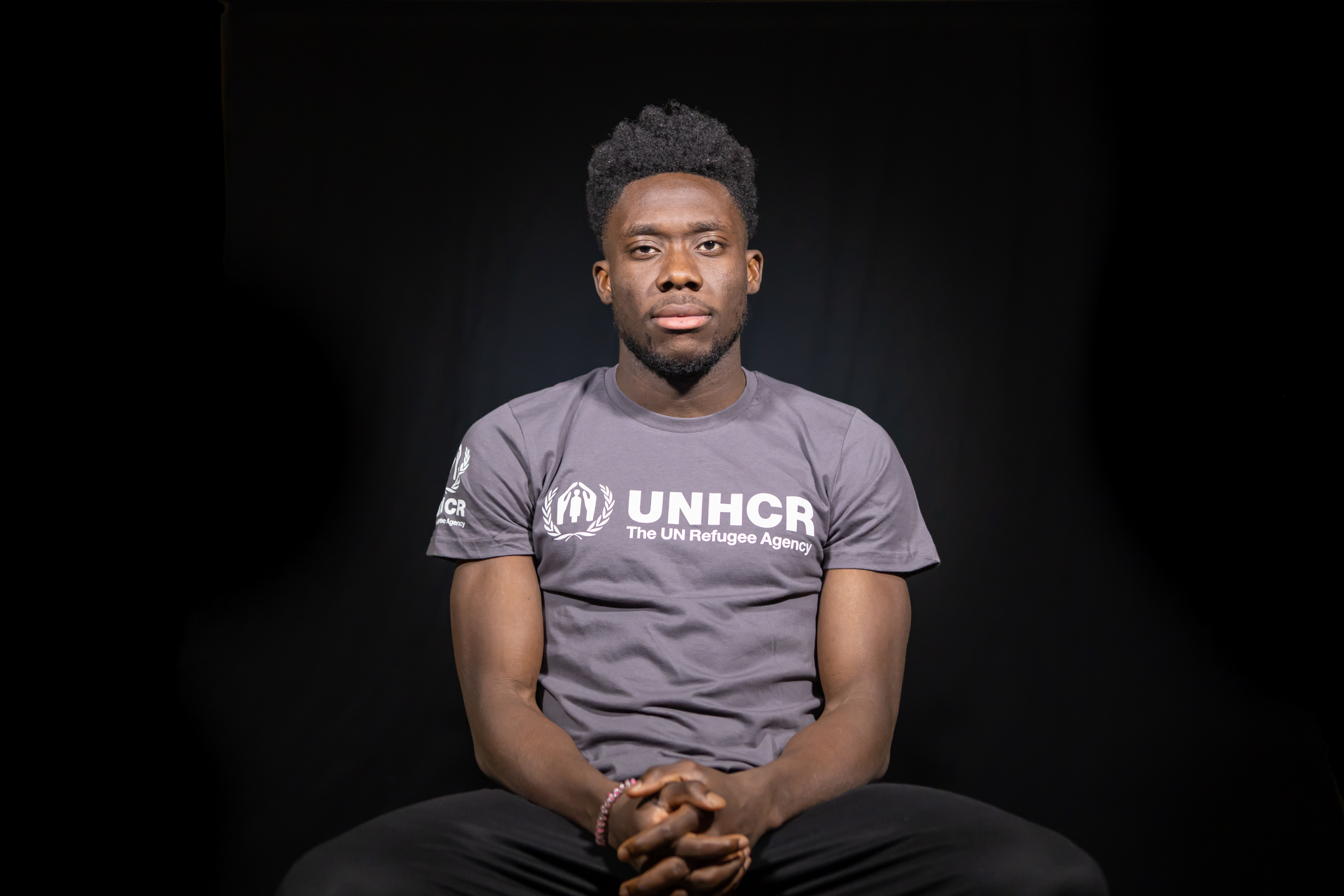 Alphonso Davies sitting in chair, wearing UNHCR t-shirt