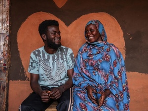 A woman and a man sit together, smiling at each other, in front of a heart-shaped wall painting