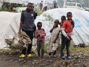 A man stands in front of UNHCR tents, with a young child on his back and four other children standing beside him