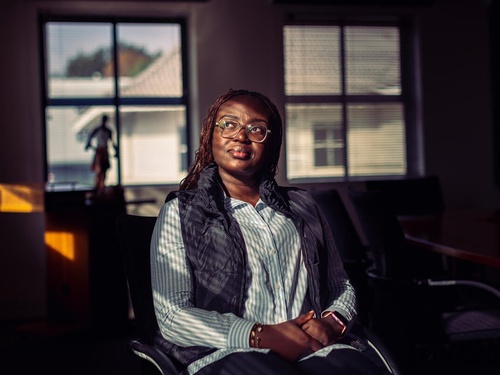 A woman sitting indoors in dappled light, with windows behind her, gazes into the distance