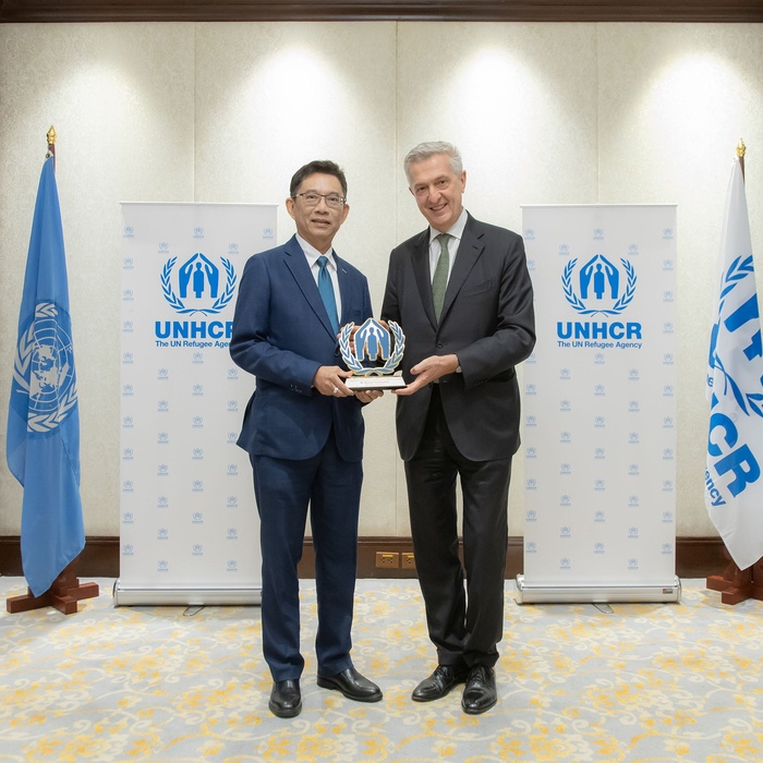 Mr. Wanchai Tachavejnukul and Filippo Grandi smiling and holding an award together, standing in front of UNHCR banners and flags