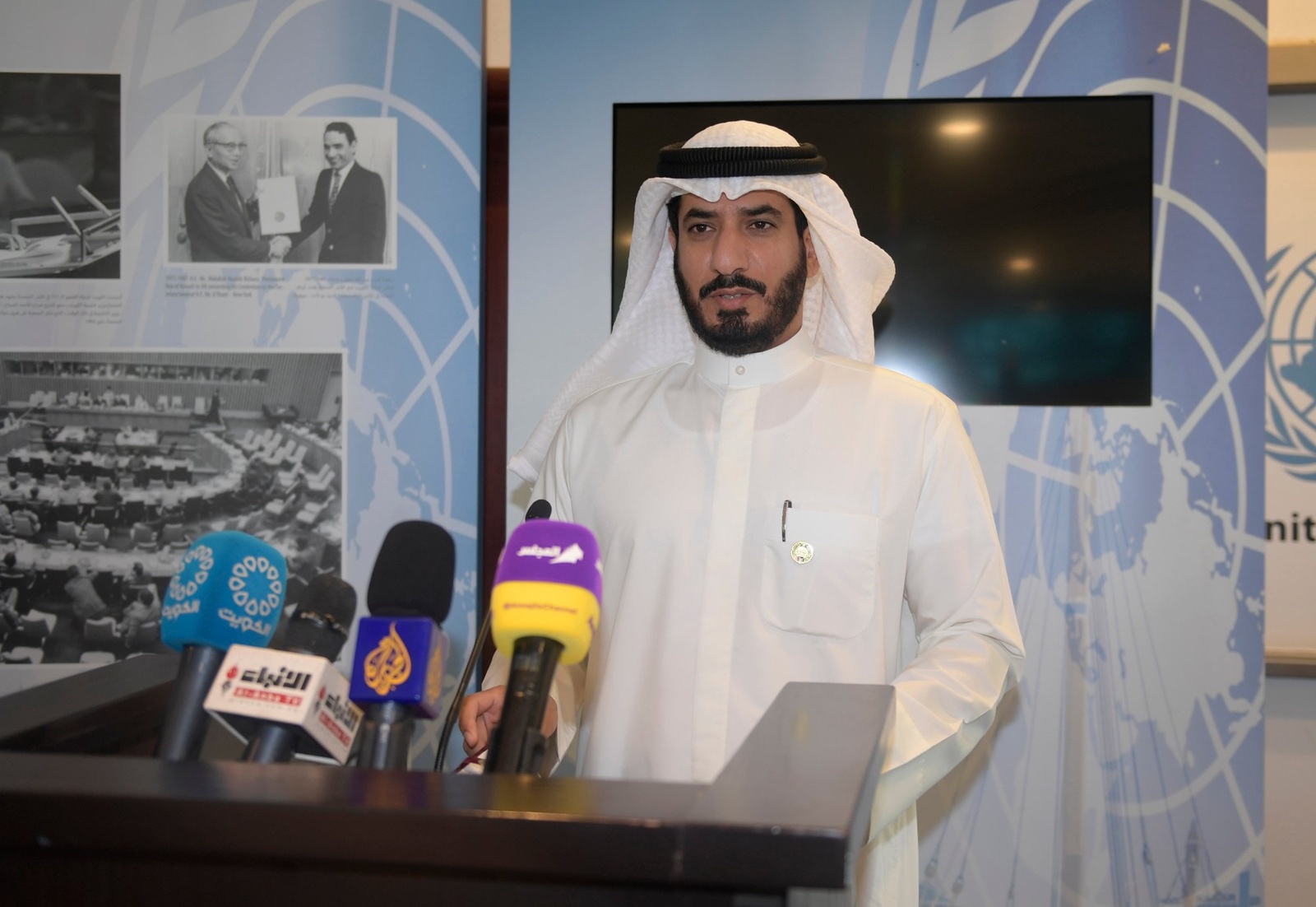 Mr. Majed AlAzmi at a lectern with many microphones. There is a blank TV and UNHCR historic documents behind him.