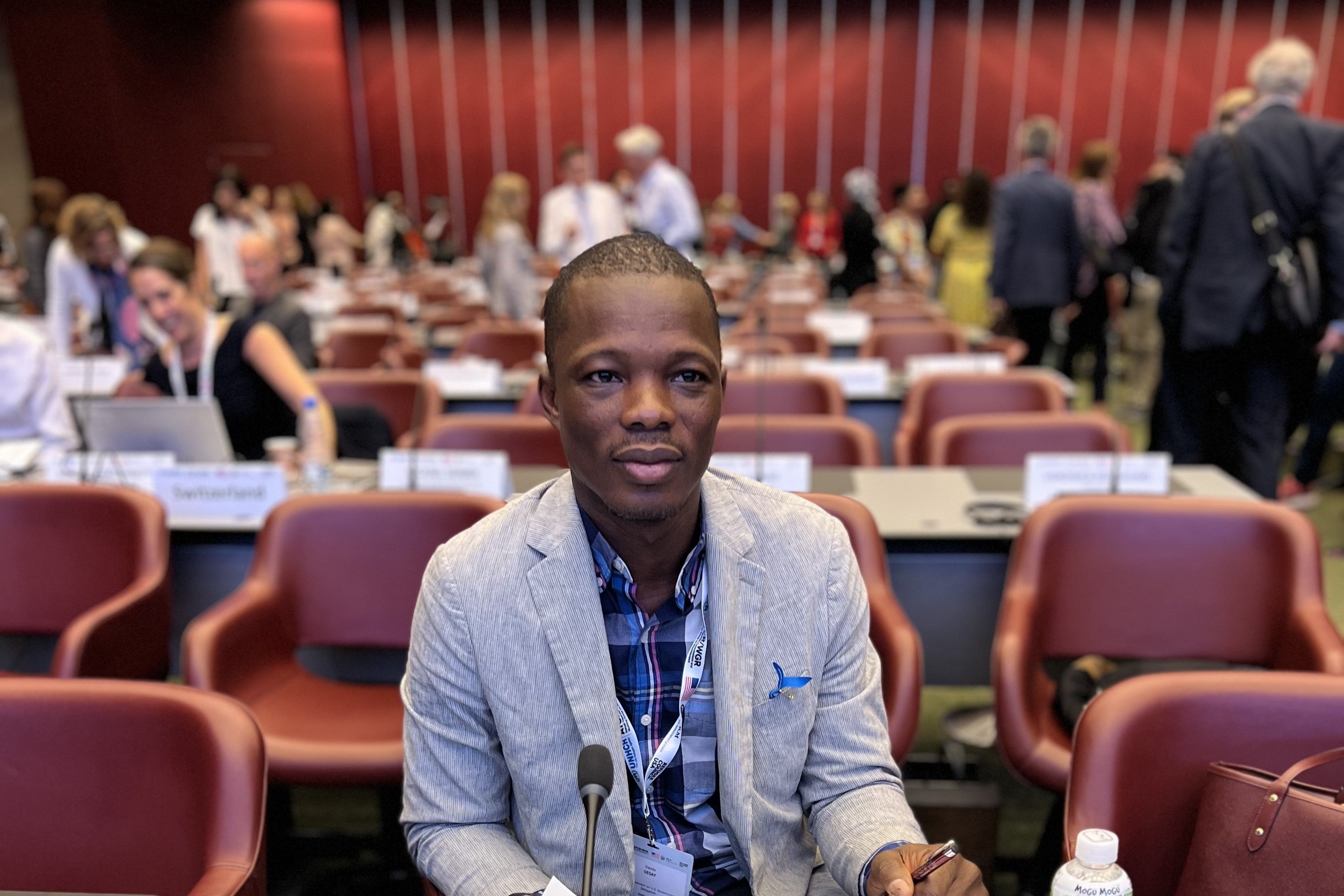 A man sits in a conference room with other delegates in the background.