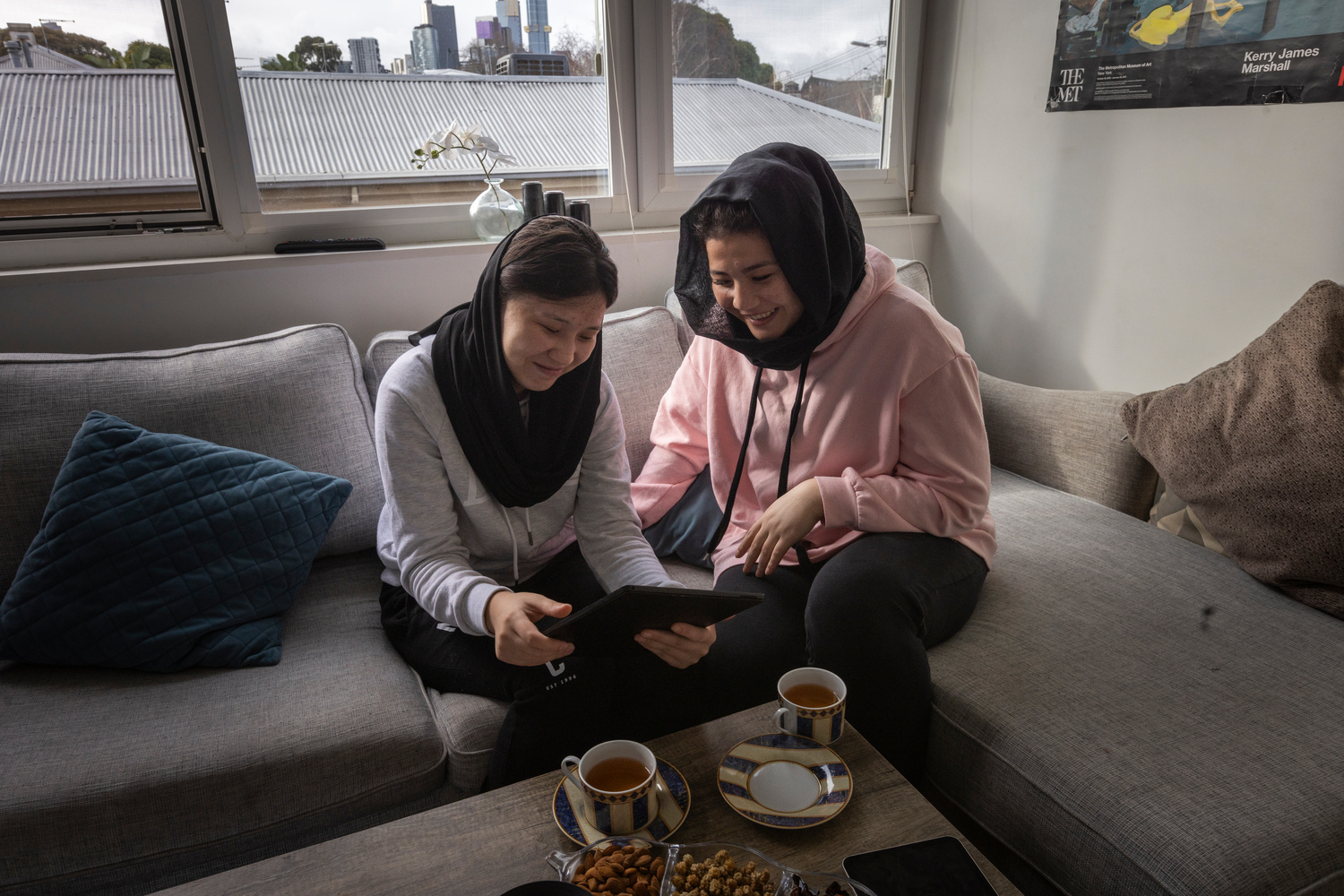 Two young women sit on a couch together looking at a tablet and drinking tea.