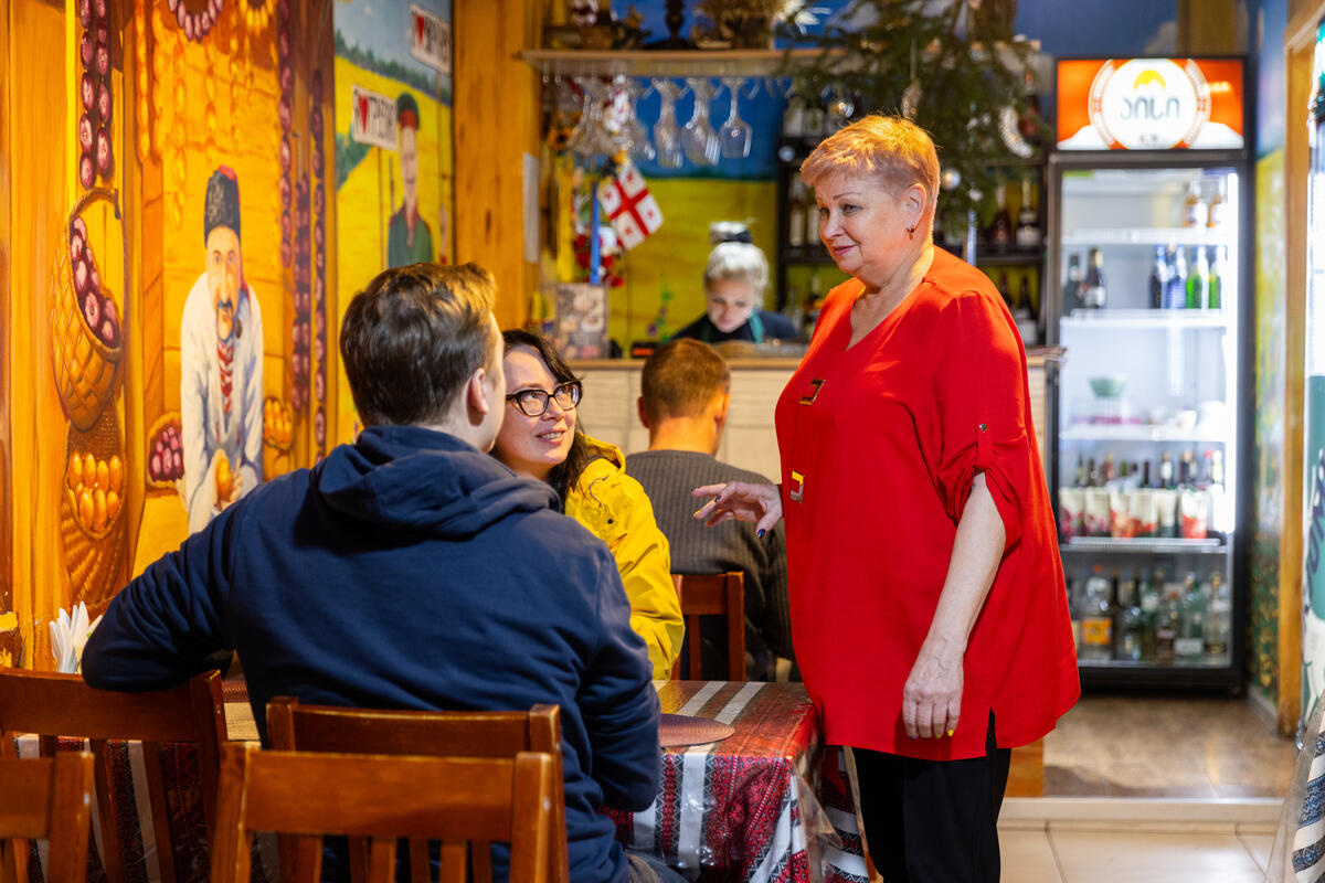 A smiling woman in red serves customers inside a colorful café